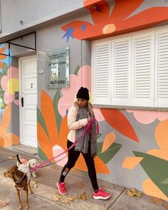 a woman walking her dog on a leash in front of a colorful wall with flowers