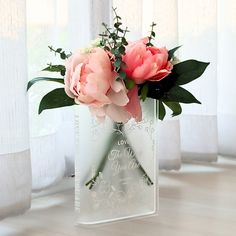 two pink flowers in a clear vase with greenery on the table next to white curtains