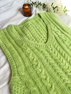 a green knitted sweater sitting on top of a bed next to a vase with flowers