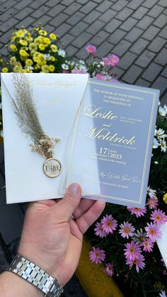 a person holding two wedding cards in front of some flowers and potted plants on the sidewalk