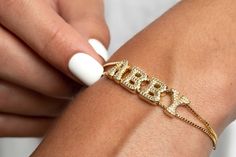 "Introduce our First Retro Gold Pavé Custom Slider Bracelet, and it's absolutely breathtaking.  This bracelet boasts two strands of box chain and letter beads that you can personalize with your name, initials, or favorite phrase.  The Charms are designed in a retro bubble letter font and feature intricate pavé detail, which gives it a luxurious touch.  Moreover, the pull-tie closure makes it simple to adjust to your preference.  You can wear this bracelet every day, and it'll undoubtedly become a staple in your jewelry collection.  Make sure to explore the Letter Key to see all the options for customization! * Material: High-quality and Anti Tarnish 14K Gold Plated - Brass; AAA Grade Cubic Zirconias. * Finish: Up to 5.75\" (14.5 cm) in diameter, adjustable 14K Gold Plated Box Chain.  HOW T Name Bracelet Gold, Bubble Letter Fonts, Bracelet Name, Gold Bubbles, Bubble Letter, Slider Bracelet, Dope Jewelry, Gold Bracelet For Women, Funky Jewelry