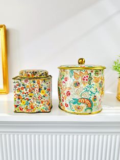 two decorative canisters sitting on top of a white mantle next to a potted plant