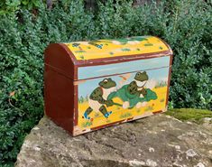 a painted wooden box sitting on top of a rock next to some bushes and trees