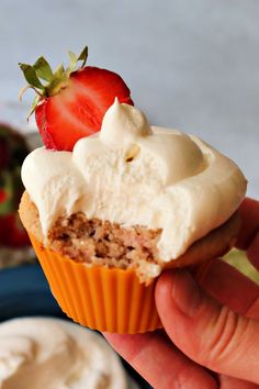 a hand holding a cupcake with cream frosting and a strawberry on the top