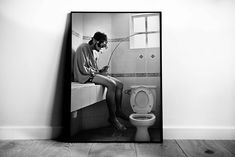 a black and white photo of a man sitting on a toilet looking at his cell phone