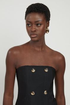 a woman in a black dress with gold buttons on the front and side, looking at the camera