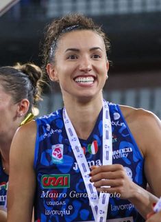 a smiling woman holding a medal in her hand and wearing a blue shirt with white ribbons around her neck