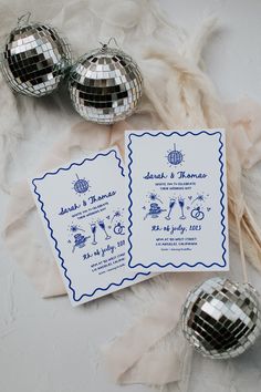 two silver disco balls and some cards on a white table cloth with feathers in the background