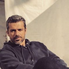 a man sitting on top of a chair next to a white wall and wearing a black hoodie