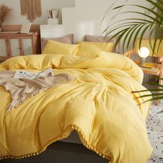 a yellow comforter with pom poms on the bottom is in front of a potted plant