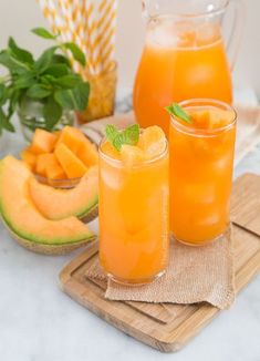 two glasses filled with orange juice sitting on top of a cutting board next to sliced melon