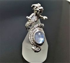 Dragon Ring with Natural Moonstone Pure .925 Sterling Silver Unisex Ring with Genuine Moonstone Gemstone This beautiful Exclusive Design Dragon Ring has been Fabulously casted with .925 Sterling Silver The weight of the Ring is 12.5 grams The Ring Size is Adjustable from size 5 to size 10 *please inquire if you don't see your size listed* Please allow 2-3 weeks shipping as we are in Bangkok Thailand at this time We are a Los Angeles based company operating out of Thailand so that we may bring yo Mystical Silver Crystal Ring With Gemstone, Silver Crystal Ring With Gemstone, Mystical Moonstone Jewelry With Large Stone, Celestial Gemstone Jewelry Collectible, Collectible Celestial Gemstone Jewelry, Mystical Sterling Silver Jewelry, Mystical Silver Round Jewelry, Mystical Silver Jewelry With Large Stone, Mystical Round Silver Jewelry