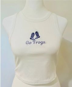 Introducing the Go Frogs Cropped Tank: A chic white cropped tank top crafted from 97% polyester and 3% spandex. Featuring a light and dark purple embroidered cowgirl boot design, embroidered below the cowgirl boot is Go Frogs in a charming orchid cursive font. Western Style Fitted Sleeveless Tank Top, Fitted Sleeveless Western Tank Top, Fitted Western Style Sleeveless Tank Top, Fitted Western Tank Top For Summer, White Cropped Tank Top, Frog Tank, Womens Tank Tops, Boot Design, White Crop Tank