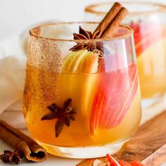 two glasses filled with apple cider sitting on top of a cutting board next to cinnamon sticks