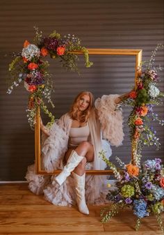 a woman sitting in front of a frame with flowers and feathers around her legs, posing for the camera