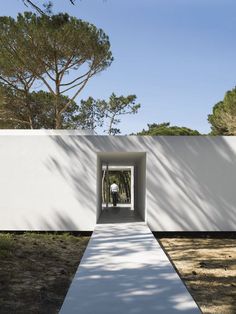 an empty walkway leading to a white wall with trees in the background and a man standing at the end