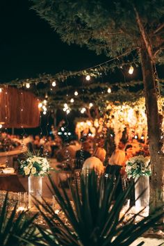 people are sitting at tables with lights strung from the trees and plants in vases