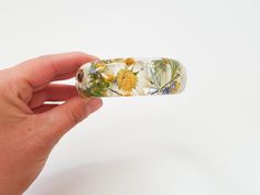 "Real flower resin bracelet. The bracelet features real pressed Daises, ferns, cornflowers and yellow baby's breath flowers. The bracelet is made with jeweler's grade resin. Available in 3 different sizes: Small: diameter/opening 2.4\" (63mm) , height of the bracelet is approximately 1\" (25mm) Medium: diameter/opening 2.6\" (66mm) , height of the bracelet is 1\" (25mm) Large: diameter/opening 2.72\" (69mm) , height of the bracelet is 1\" (25mm) XS and XL sizes available upon request If you are unsure if it will fit you, please feel free to contact us and we will send you instructions on how to measure your hand and see if this bangle size will fit. PERSONALIZATION ADD-ON IS AVAILABLE FOR THIS BRACELET If you would like a personalized engraved plate on this bracelet, you must purchase the Nature Gifts, Pressed Flower Jewelry, Breath Flowers, Jewelry Nature, Flower Daisy, Resin Bracelet, Daisy Bracelet, Resin Bangles, Flower Resin