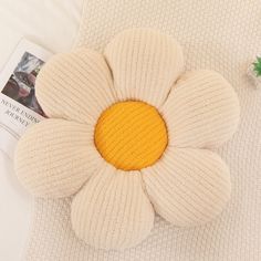 a knitted flower pillow sitting on top of a bed next to a magazine and plant