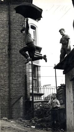 black and white photograph of boys jumping off roof with umbrellas over their heads in an alleyway