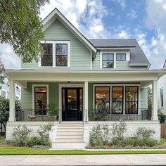 a green house with two story windows and white trim on the front porch is surrounded by greenery