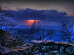 the sun shines through clouds over trees on top of a rocky cliff in the mountains