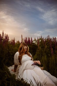 a woman in a white dress is sitting on the ground with purple flowers behind her