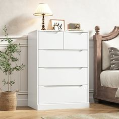 a white chest of drawers in a bedroom next to a bed and nightstand with a plant