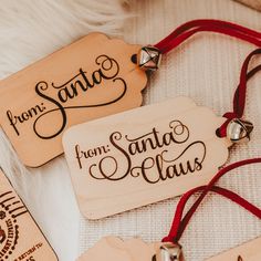 three wooden tags that say santa claus and santa feliz on the front, with bells attached to them