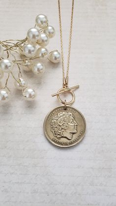 This authentic and vintage Greek coin makes a fabulous gold medallion necklace. It is a  1994 100 drachma coin set on an 18" gold-plated chain. Your choice of a front toggle closure or a back lobster clasp with a 3" chain extender. The coin has been polished and matches the chain beautifully. Welcome to Recycled Finery!  I repurpose old jewelry, coins, beads, vintage buttons, watch parts, and charms into new jewelry.  My ever-growing collection of these materials comes from estate sales, rummage sales, antique and thrift stores, and family and friends' collections they no longer need or want. I have fun putting these new pieces together and hope you will find them fun to wear and as unique gifts for your loved ones.     Please note: Because my components are recycled from old or vintage je Antique Gold Medallion Necklace For Gift, Nickel-free Yellow Gold Medallion Coin Necklace, Classic Coin Pendant Necklace For Commemoration, Brass Medallion Coin Necklace For Gift, Vintage Tarnish Resistant Medallion Necklace, Classic Gold Medallion Necklace With Coin Pendant, Vintage Brass Coin Necklace With Coin Pendant, Antique Round Coin Necklace, Vintage Brass Coin Pendant Necklace