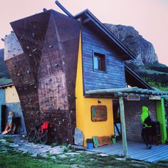 a person standing in front of a house made out of rocks and wood with mountains in the background