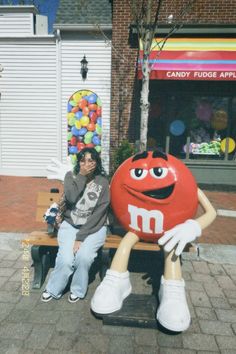 a woman sitting on a bench next to a giant m & m ballon in front of a store