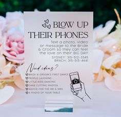 a close up of a sign on a table with flowers in the background and someone holding a cell phone