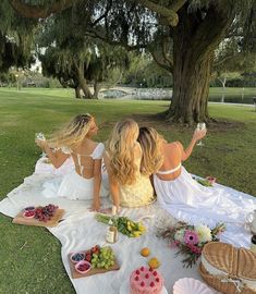 two women sitting on a blanket in the grass with plates of food and drinks next to them