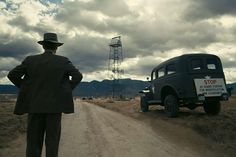 a man in a suit and hat standing on a dirt road next to an old truck