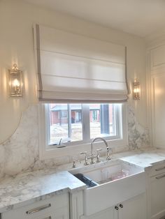 a white kitchen with marble counter tops and sink under a window that has roman shades on the windowsill