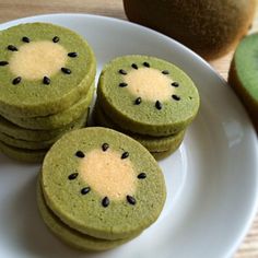 four green cookies with black seeds are on a white plate next to an kiwi