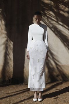 a woman standing in front of a wall wearing a white dress with flowers on it