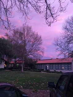 cars parked in front of a building with trees on the other side and pink sky