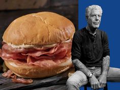 an older man sitting on a bench next to a large sandwich with meat in it