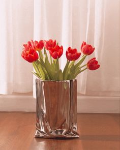 red flowers in a silver vase on a wooden floor near a window with white curtains