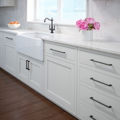 a kitchen with white cabinets and wooden floors, pink flowers in a vase on the window sill