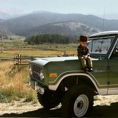 a young boy sitting on the hood of a green truck with mountains in the background