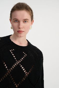 a woman with blue eyes wearing a black sweater and gold earrings, standing in front of a white background