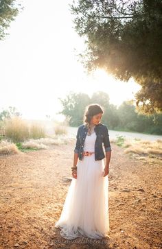 a woman in a white dress and jean jacket is standing on the dirt road with her hands in her pockets