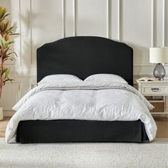 a bed with white linens and black headboard next to a plant in a vase