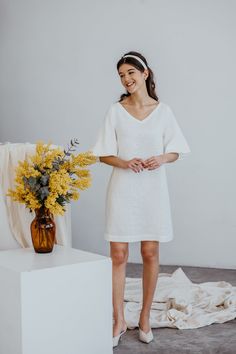 a woman standing next to a vase with yellow flowers in it and wearing a white dress