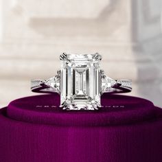 an emerald - cut diamond ring sits atop a purple velvet covered stand in front of a white background