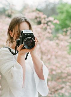 a woman holding up a camera in front of her face with pink flowers behind her