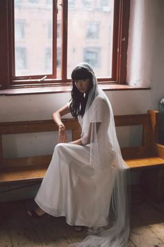 a woman sitting on a bench in front of a window wearing a white dress and veil